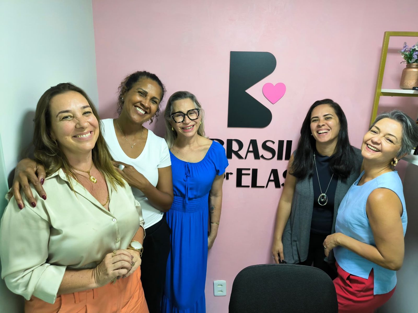 Cinco mulheres sorrindo diante do logotipo da Brasil por Elas na parede rosa
